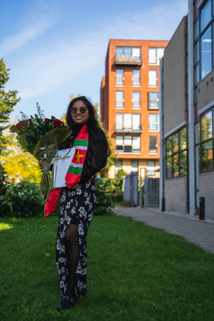 Fariel Soeleiman heeft een groot boeket rozen in haar rechterhand, haar HKU-diploma in linkerhand, een sash met de Surinaamse vlag erop om haar nek en een lange bloemetjes rok met een split aan. Ze staat buiten voor de HKU.