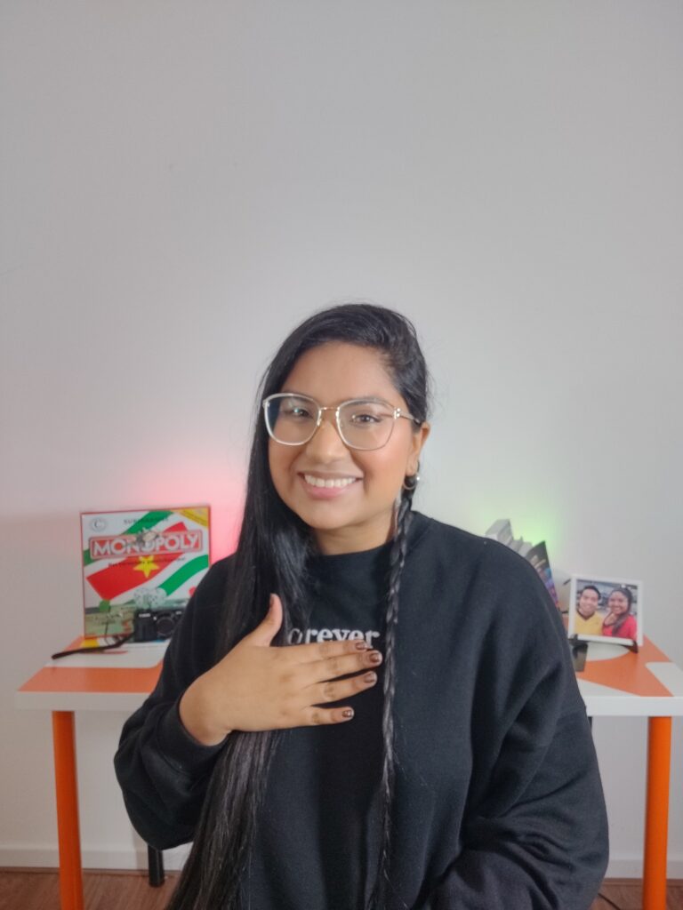 Portretfoto van Fariel Soeleiman met in de achtergrond een bureau waarop de Surinaamse Monopoly staat, een aantal boeken op een boekensteun. en een foto van haar en Deon. Ze heeft een zwarte sweater aan met het opschrift forever.