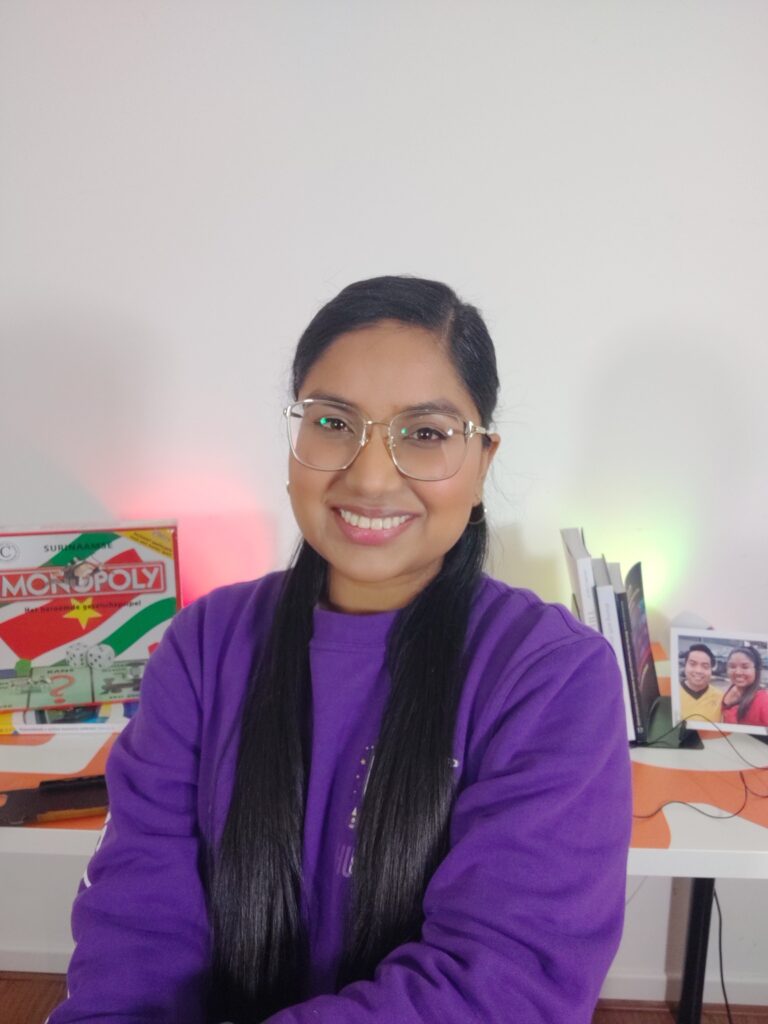 Profielfoto van Fariel Soeleiman met in de achtergrond een bureau met de Surinaamse Monopoly, een foto van haar en Deon en een aantal boeken op een boekensteun. Ze heeft een paarse sweater aan.