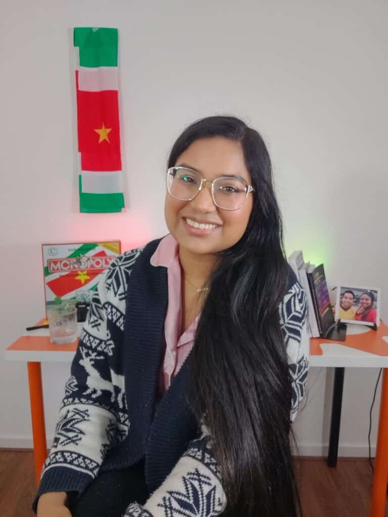 Portretfoto van Fariel Soeleiman met in de achtergrond een bureau met de Surinaamse Monopoly, een foto van haar en Deon en een Suriname sash die hangt aan de muur.