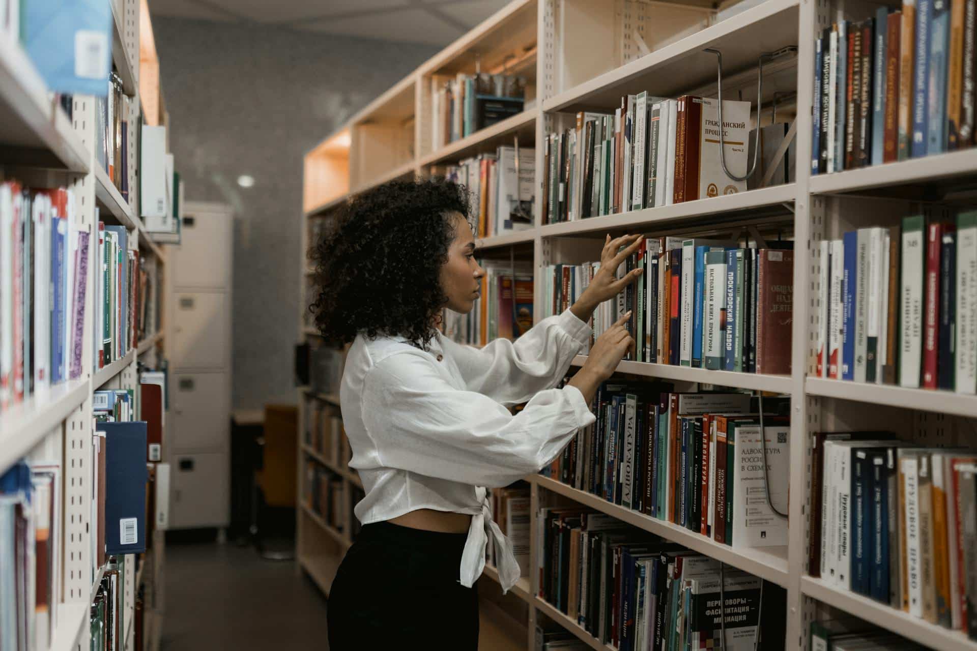 Een jongedame met een grote bos krullen zoekt aandachtig door een boekenkast in een bibliotheek.