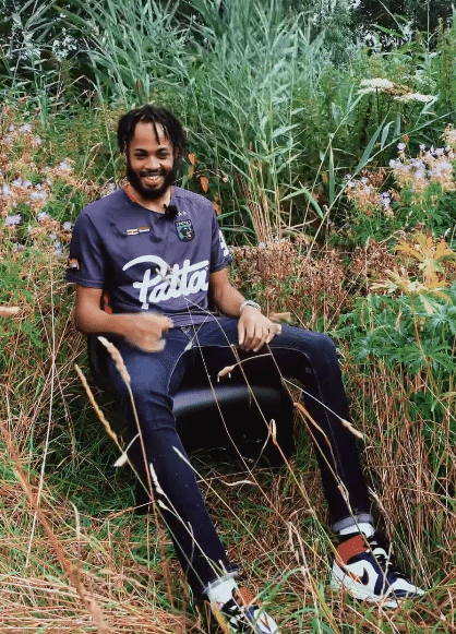 Een jongen met korte dreadlocks en een volle zwarte baard zit lachend op een stoel buiten tussen de bloemen en hoge grasstengels.
