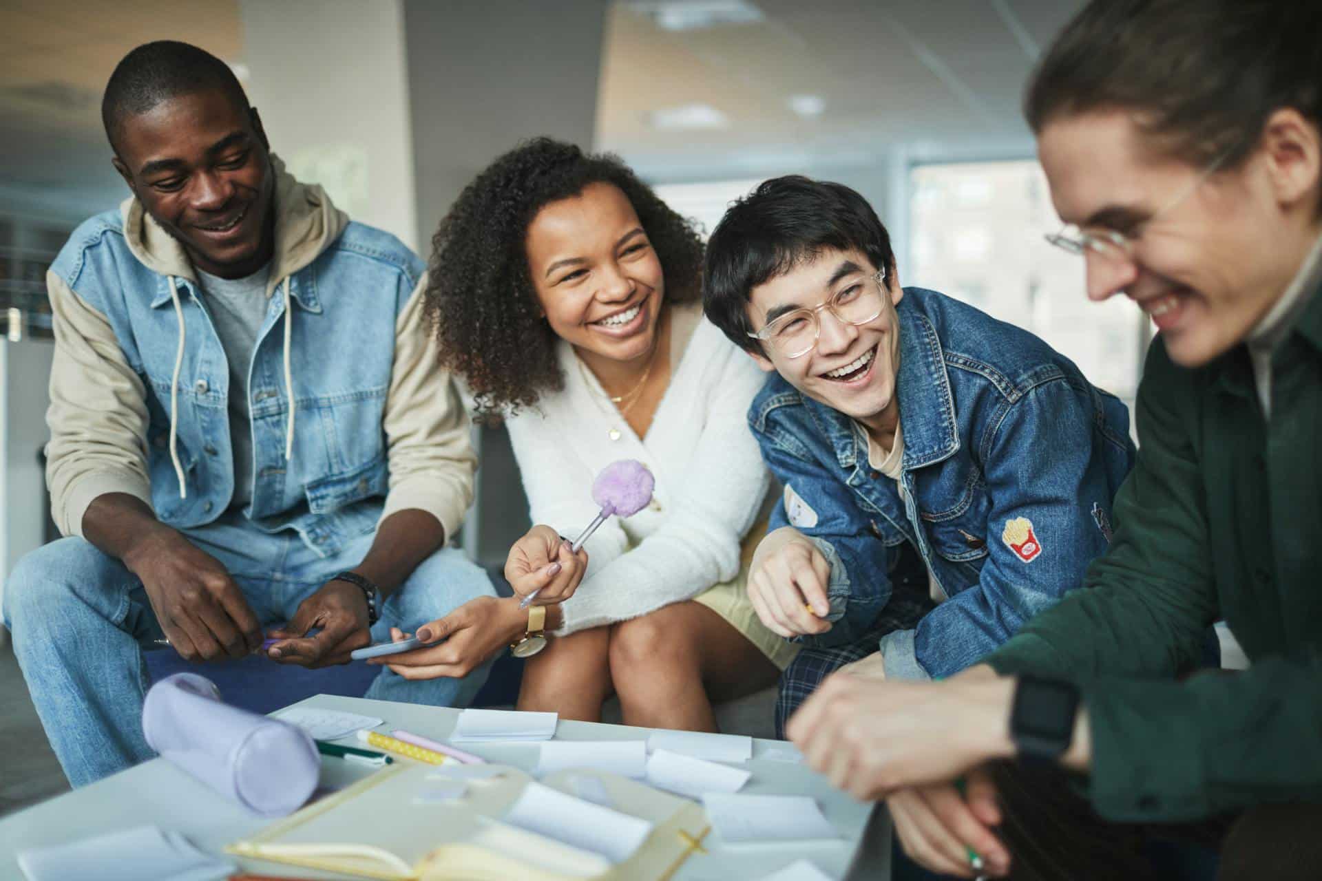 Een groep jongvolwassenen zitten in een groep aan een tafel vol notities en lachen hartelijk naar elkaar toe.